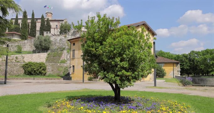 Castello di Brescia fortress and flag tower in Brescia Italy, view from the square shows sunlit stone walls cypress trees and a small park lawn as flags flutter on the keep