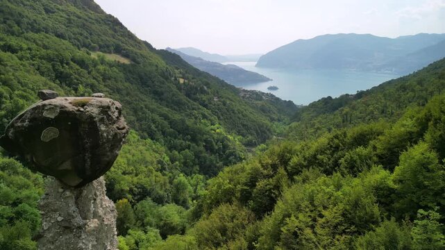 Drone flies between Zone earth pyramids with boulder caps, rising above a forested gorge toward Lake Iseo in Lombardy Italy with distant mountains