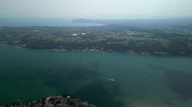Drone pullback with gimbal tilt over Lake Garda near Salo, flying backward as the camera lowers from open water to the curved promenade and terracotta old town roofs with ferry pier