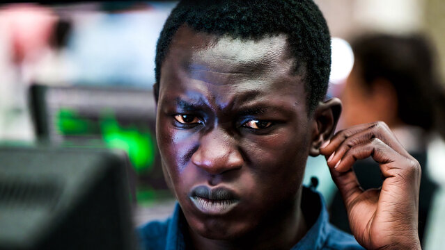 A young Black man intently stares at a computer screen, furrowing his brow in concentration. He wears a blue shirt.