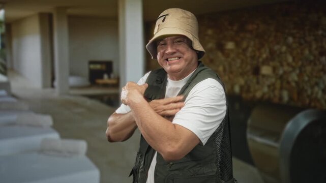 Man hugging shoulders and grimacing in a building pool area while wearing a fishing vest and bucket hat; discomfort solitude.