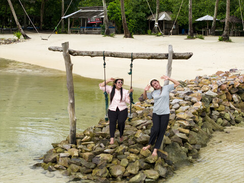Aerial view of two women enjoying a swing along Bobby Beach, where turquoise waters meet the sandy shore under the shade of swaying palms, Karimunjawa, Central Java, Indonesia.