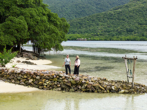 Aerial view of two figures stand on a rocky outcrop, framed by verdant trees against the tranquil waters of Bobby Beach, Karimunjawa, Central Java, Indonesia.