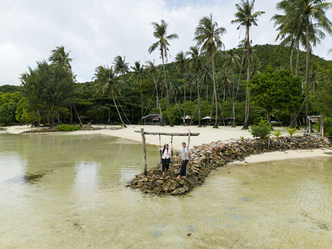 Aerial view of a couple standing on a rock formation under a wooden swing, framed by palm trees and the serene waters of Bobby Beach, Karimunjawa, Central Java, Indonesia.