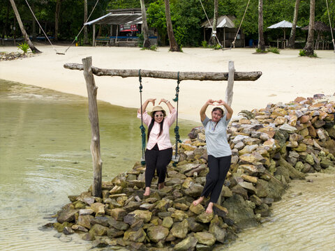 Aerial view of two women playfully pose on a swing set amidst the rocky shore, framed by swaying palm trees and tranquil waters at Bobby Beach, Karimunjawa, Central Java, Indonesia.