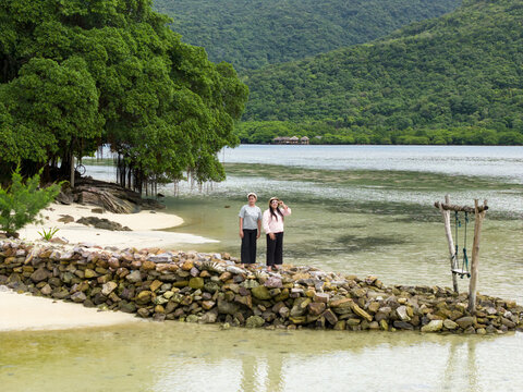 Aerial view of two figures stand on a rocky breakwater, gazing out at the shimmering turquoise water and lush green hills, Bobby Beach, Karimunjawa, Central Java, Indonesia.