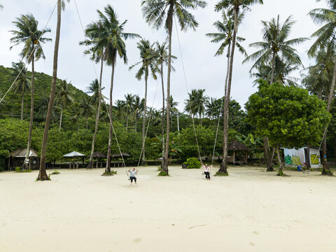 Aerial view of Bobby Beach with its pristine white sand meeting the lush green forest, with people playing on swings hanging from palm trees, Karimunjawa, Central Java, Indonesia.