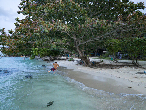 Aerial view of a person on a swing under a lush tree at Tanjung Gelam Beach, where the turquoise water meets the sandy shore, Karimunjawa, Central Java, Indonesia.
