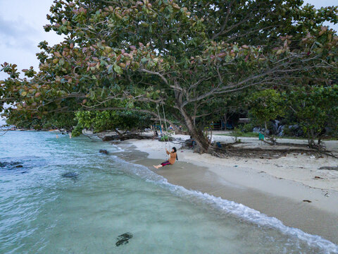 Aerial view of a lone figure on a swing beneath a sprawling tree on Tanjung Gelam Beach as gentle waves kiss the shore, Karimunjawa, Central Java, Indonesia.