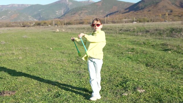 Juggling girl performs diablo stick routine outdoors on green field with majestic mountains in the background.
