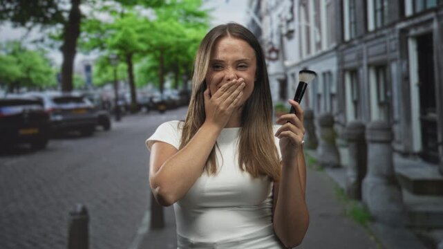 Young brunette woman holding makeup brush covers mouth with hand while smiling on a cobblestone city street lined with row houses; surprise playfulness.
