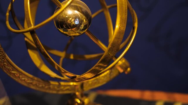 Detailed close up of a brass armillary sphere astronomical instrument against a dark blue wall. The camera performs a smooth tilt down revealing golden metallic textures and ancient design.