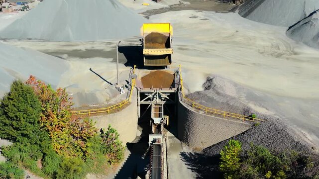 Dump truck positioned above hopper emptying and filling it with red aggregate in quarry, aerial approach
