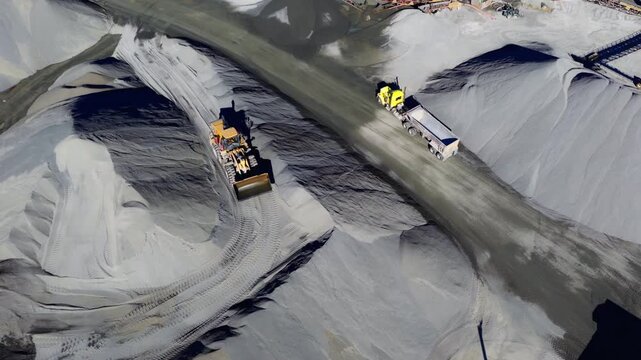 Dump truck driving between gravel piles as front loader backs up on aggregate, pullback aerial reveal