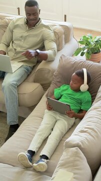 Vertical video: Reaching dad gesturing engaging son on sofa back at laptop as son on tablet green T