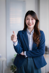 Young Asian businesswoman in a blue blazer smiling and giving a thumbs-up in a corporate studio, conveying confidence, success, approval and a positive professional attitude
