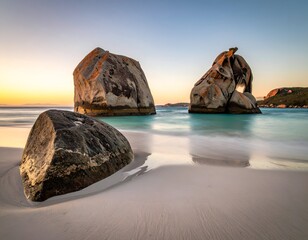 Coastal scene with large granite boulders in calm, turquoise water at sunrise. Smooth sands and distant hills