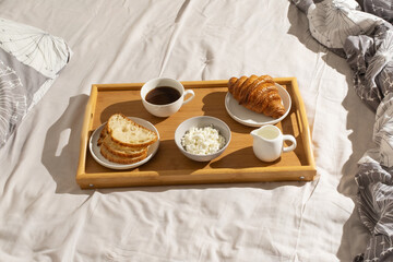 Breakfast tray with coffee, bread, cheese, and croissant on a bed during morning hours
