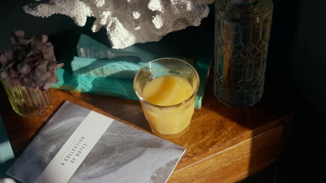 Sunlight glows softly on wooden table with glass of juice. Decorative coral, vase, and book rest beside glass. Mood is calm and cozy, perfect for relaxation or morning scenes