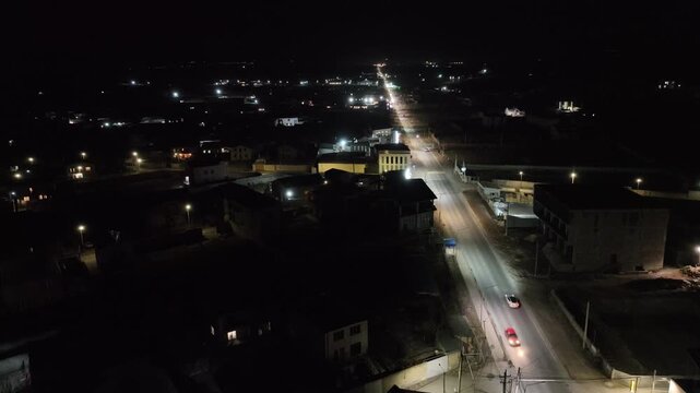 Night aerial view of quiet town, illuminated streets, scattered homes, calm atmosphere. Streetlights glow along main road, vehicles move slowly. Ideal for urban planning, travel. No people visible