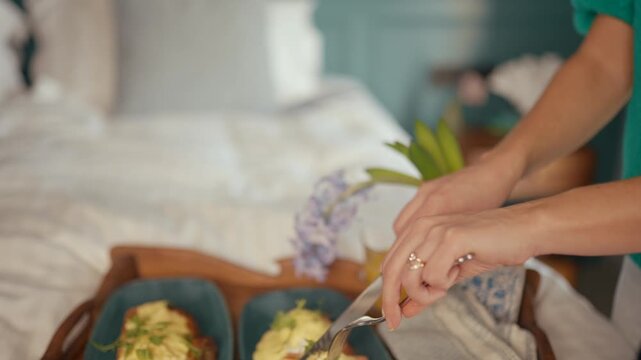 Person serves breakfast in bed. Hands cut eggs benedict on a tray. Purple flowers and orange juice add freshness. Soft morning light warms the scene. Ideal for lifestyle, wellness