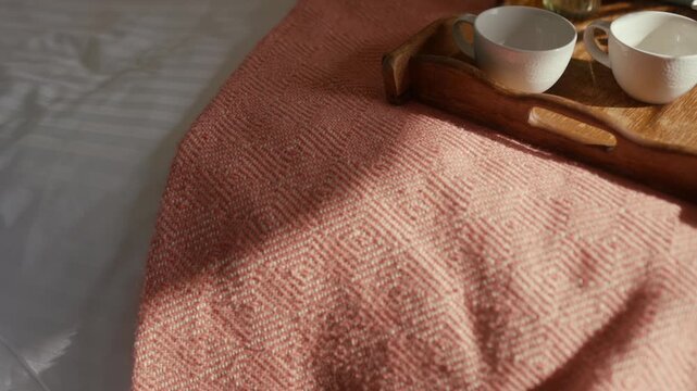 Soft light falls on cozy bed with pink textured blanket. Wooden tray holds white teacups and milk jug. Hand pours tea into cup with gentle care. Scene evokes calm morning ritual or quiet relaxation
