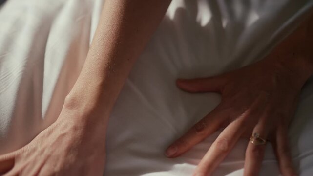 Soft, wrinkled white bed sheets fill the frame. Hands rest gently on the bedding, relaxed and calm. Ring glints on one finger, hinting at intimacy. Ideal for scenes of rest, romance