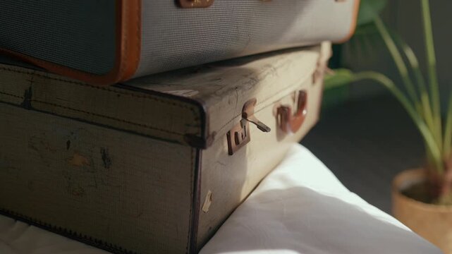 Hand gently touches vintage suitcases stacked on bed. Warm sunlight highlights textured leather and metal clasps. Scene suggests travel memories or upcoming journey