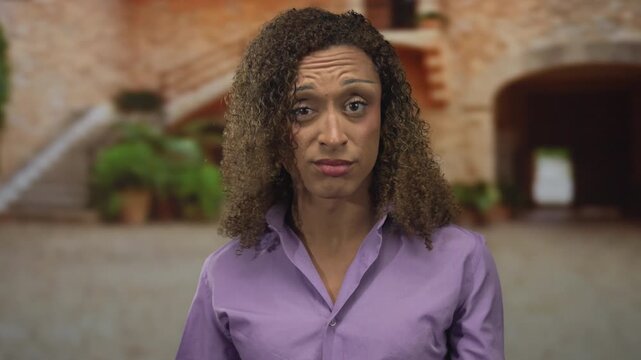 Woman with curly hair holds her fingers in air quotes while standing in an old town street with a furrowed brow; skepticism.