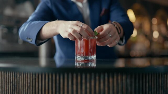 Barkeeper garnishes cocktail with mint leaf. Red drink sits on dark bar. Hands focus on finishing touch. Warm, blurred lights suggest upscale setting. Perfect for advertising or lifestyle videos