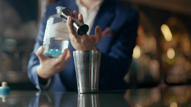 Cocktail bartender pours blue liquid into metal shaker. Hands expertly measure spirits with jigger. Bar background softly blurred, warm lights glow
