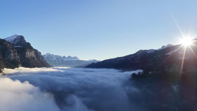 Breathtaking aerial of Alpine peaks illuminated by low winter sun. Below, a dense white fog blanket covers Lake Walensee and near Walenstadt, creating a dreamlike Swiss vista.
