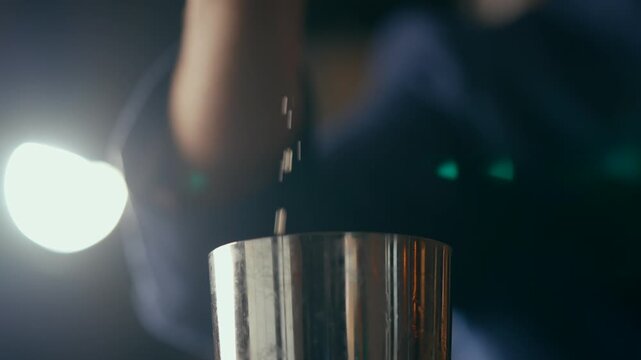 Close-up of metallic cocktail shaker, reflecting warm lights in dim bar setting. Soft bokeh background suggests nightlife ambiance. Person's hand subtly moves, implying mixing or serving drinks