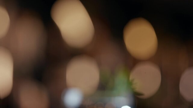 Softly blurred background with warm golden bokeh lights. Cocktail glass with clear liquid and green garnish. The glass reflects ambient lighting. Perfect for luxury, nightlife, or bar scenes