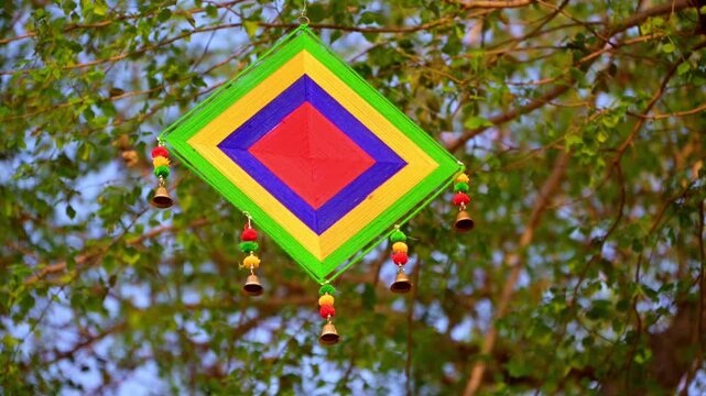 Colorful square kite hanging decoration with green, yellow, blue, and red thread patterns and small bells, gently swaying in the wind against leafy tree branches.