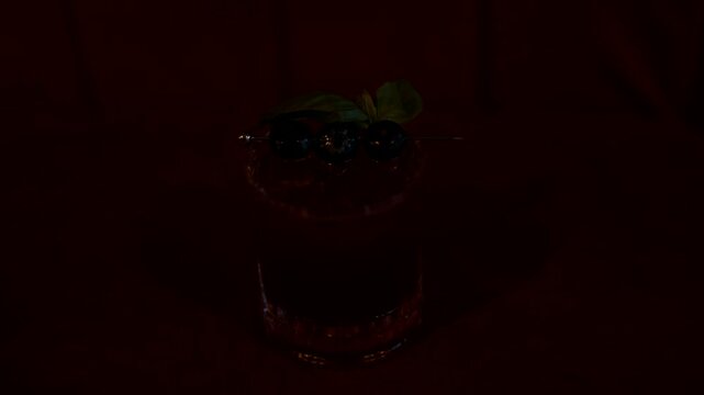 Single cocktail glass filled with pink liquid and three blueberries on a skewer. The drink is garnished with a fresh green leaf. It sits on an orange textured surface