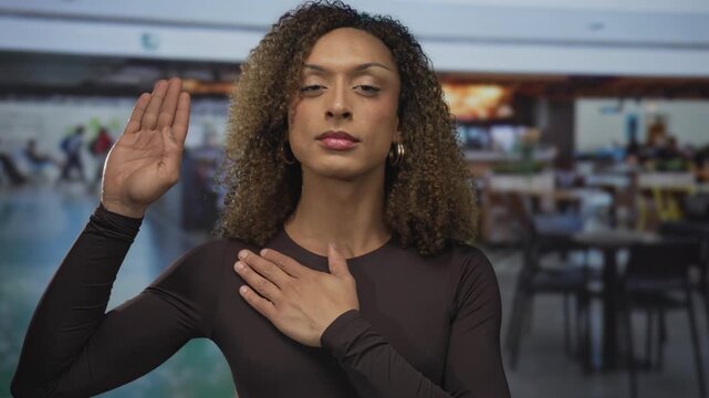 Transgender hispanic woman holds hand to chest and raises other hand inside busy airport terminal; distress.