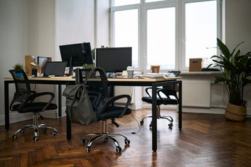 Empty modern office showing clean desks with computer monitors, office chairs neatly arranged, organized workspace with supplies and potted plant near window, no people present