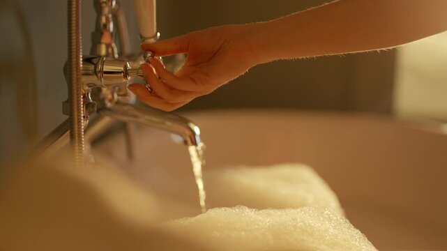 Warm water drips from vintage faucet into foamy bath. Hand adjusts tap, creating soft, rhythmic flow. Bath filled with rich bubbles, inviting relaxation. Scene evokes comfort, self-care