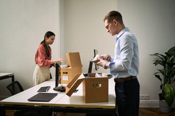 Young Caucasian man and young Asian woman cleaning office desks by packing items into cardboard boxes, both focused on organizing workspace, modern office environment visible
