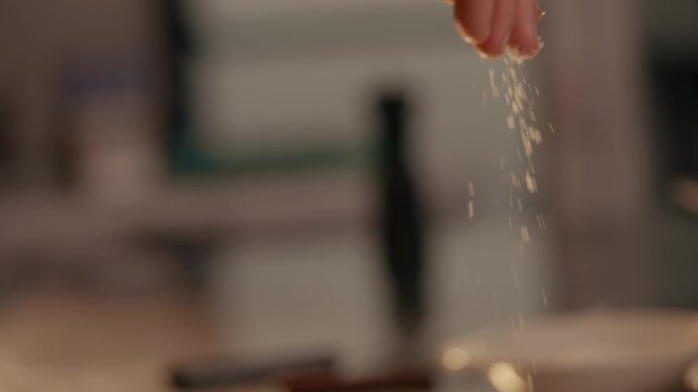 Hand sprinkles coarse salt over raw steak. Close-up captures salt falling. Kitchen background softly blurred. Action focuses on seasoning meat. Ideal for cooking, food, or restaurant content