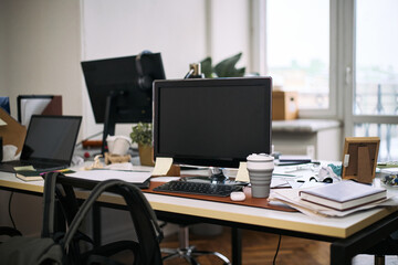 Empty office workspace showing computer monitor, keyboard, reusable coffee cup, scattered documents...