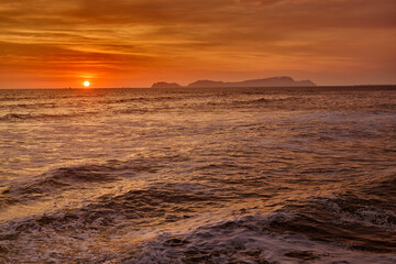 Obraz premium A vibrant summer sunset over the Pacific Ocean along Lima’s Costa Verde, with the silhouette of San Lorenzo Island on the horizon. Warm golden light reflects across the waves