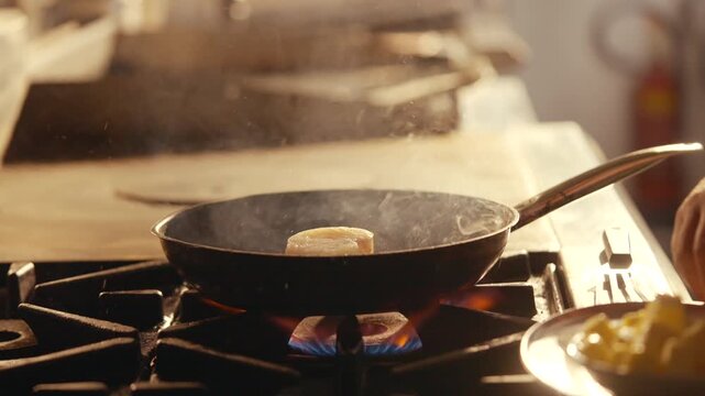 Butter cube melts in a pan over a gas stove flame. Warm kitchen light glows softly. Steam rises gently from the hot surface. Bowl of chopped vegetables sits nearby ready to add