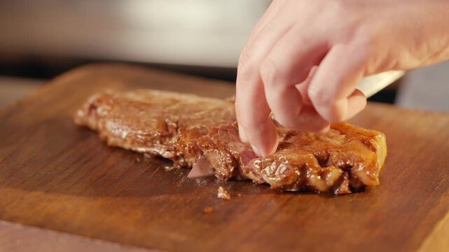 Hand slices juicy steak on wooden board. Close-up shows knife cutting meat. Warm kitchen lighting enhances food texture. Perfect for cooking, restaurant, or food-related projects