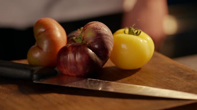 Fresh heirloom tomatoes rest on a wooden cutting board. Sharp knife lies beside them, ready for use. Warm kitchen light highlights their vibrant colors. Person's hand appears. Perfect for food blogs