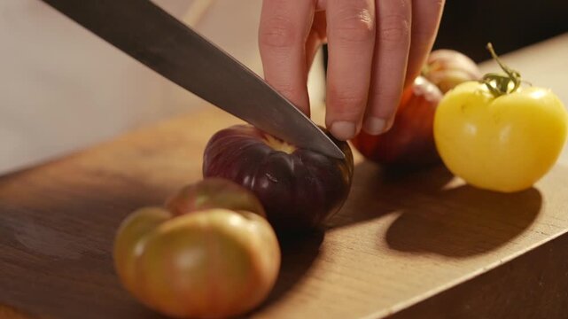 Chef slices colorful heirloom tomatoes on wooden cutting board. Close-up shows vibrant red, yellow, and purple tomatoes. Knife glides smoothly through ripe fruit, revealing juicy interior