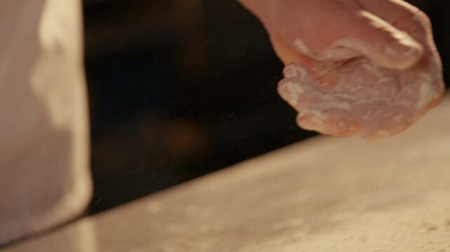 Hand sprinkles flour in warm, golden kitchen light. Dust particles float in slow motion. Scene evokes cozy baking or culinary creativity. Ideal for recipes, food blogs, or bakery branding