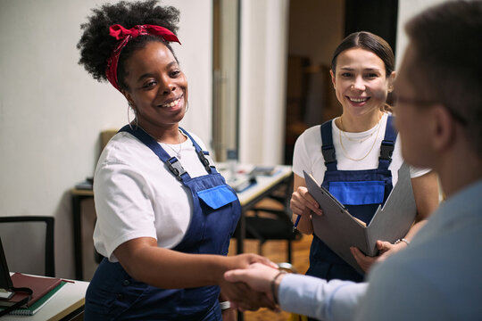 Black young adult woman and Caucasian young adult woman wearing work uniforms greeting Caucasian middle aged man in office, smiling and shaking hands, holding clipboard