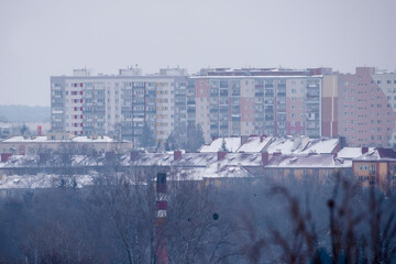 Zimowa panorama miasta (Ostrowiec Świętokrzyski) w mglisty, zimowy dzień. Panorama przedstawia mglisty dzień z dachami pokrytymi śniegiem. Na pierwszym planie widać bezlistne drzewa i komin © Grzegorz